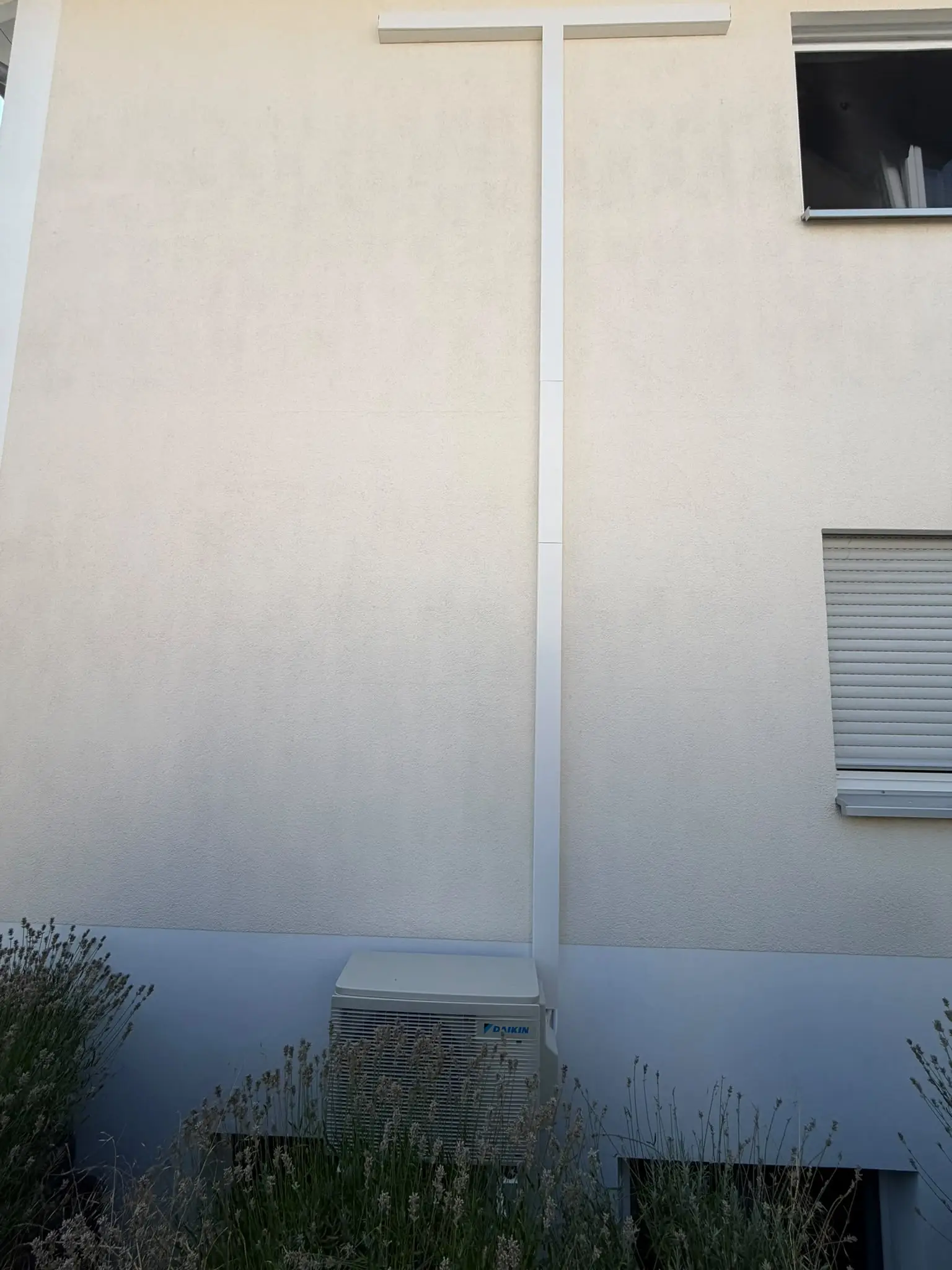 Außengerät von Daikin am Sockel einer hellen Hausfassade; ein weißer Leitungskanal führt senkrecht nach oben und waagerecht unter das Fenster.