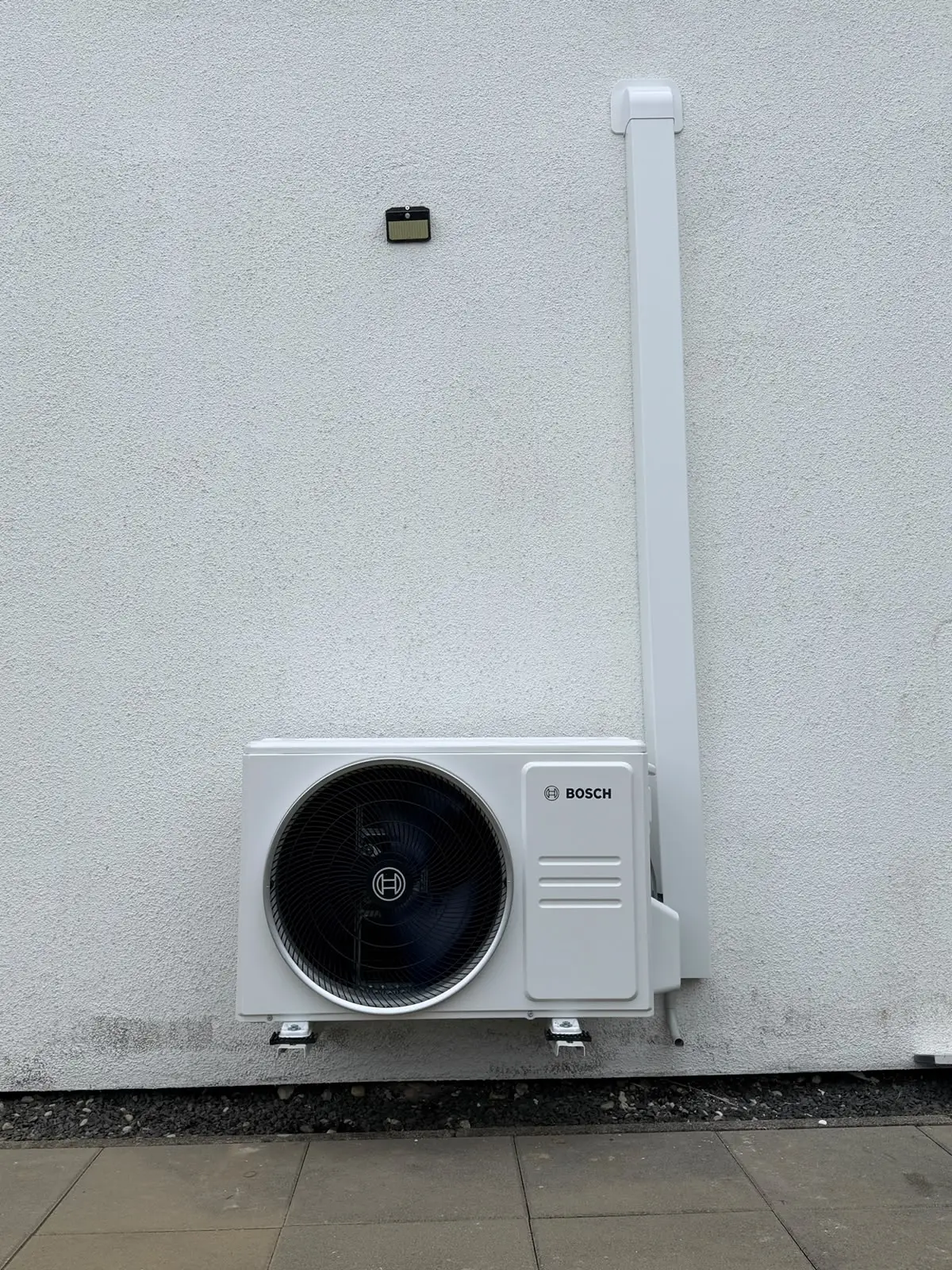 Außengerät von Bosch auf Wandkonsolen vor einer weißen Fassade, darüber ein senkrechter weißer Leitungskanal.